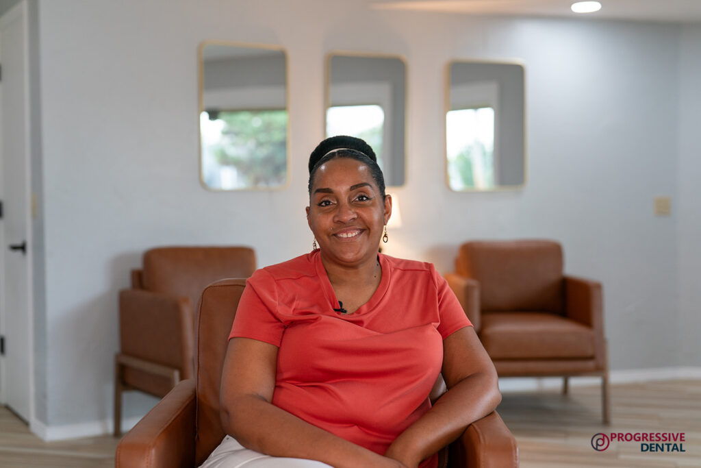 A woman in an orange shirt smiles in a modern waiting area with brown chairs and mirrors, showcasing the best cosmetic dentist in Oklahoma City. Progressive Dental logo bottom right.