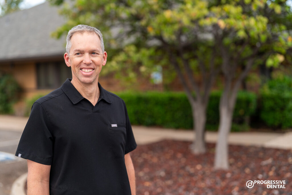 A man in a black shirt stands outside near trees and a building, smiling. “Progressive Dental”—an Oklahoma City cosmetic dentist—logo is visible in the bottom right corner.