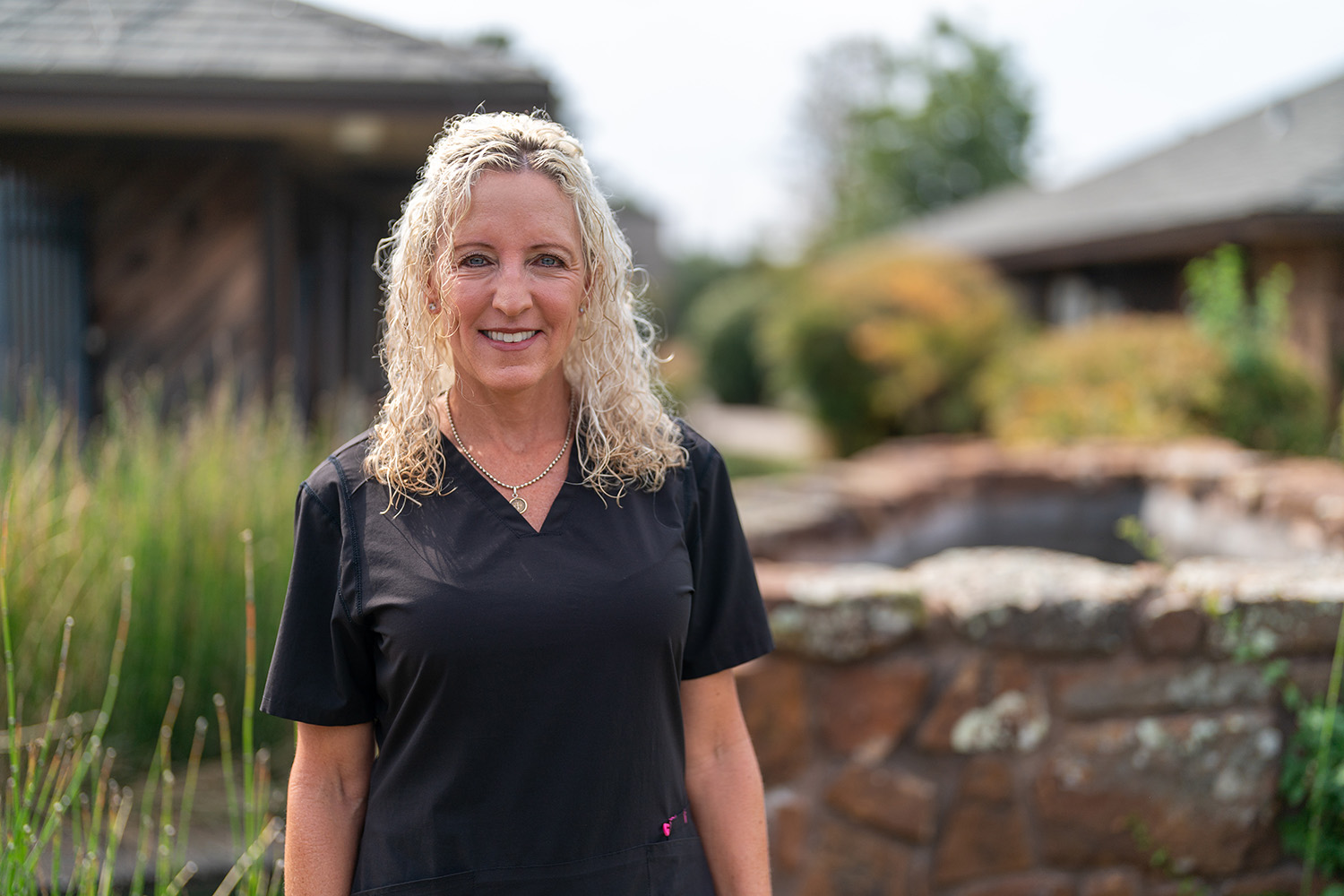 Woman with curly blonde hair wearing black scrubs stands outdoors in front of a stone wall and greenery, smiling at the camera.