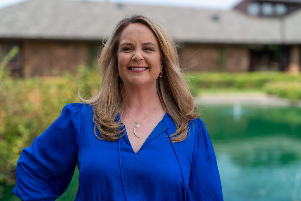 Woman with long blonde hair in a blue blouse, smiling and standing outdoors in front of a blurred building and green water.