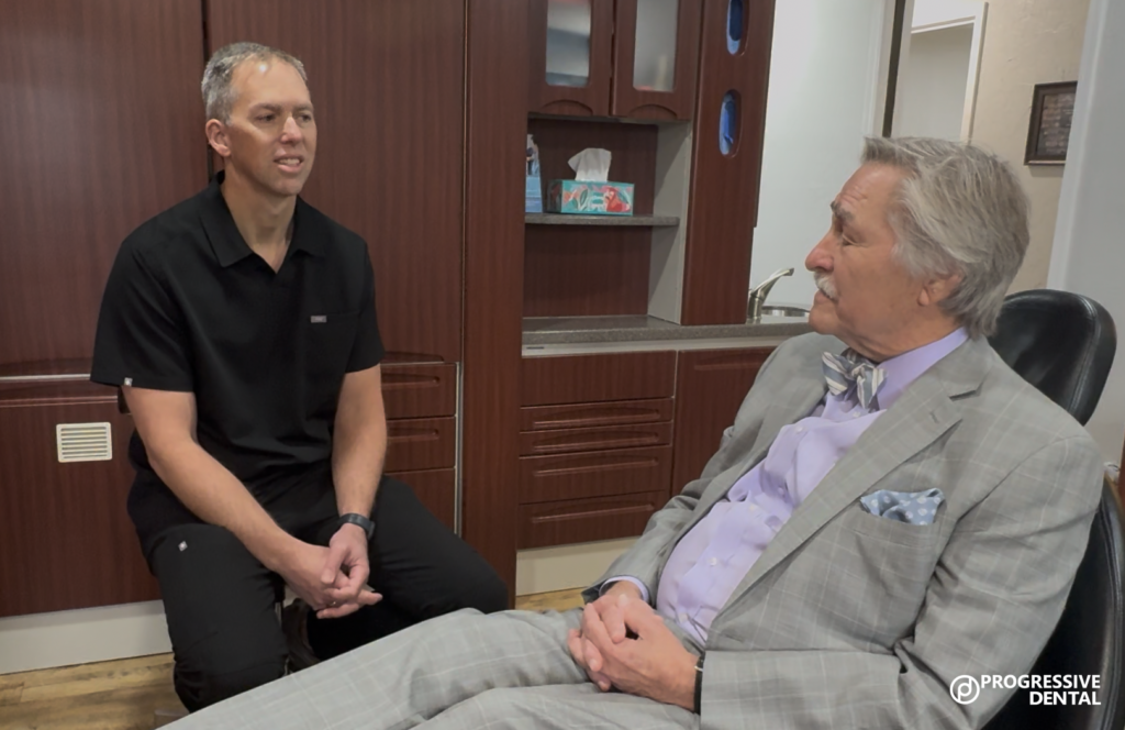 A dentist in black scrubs speaks with an older man in a suit sitting in a dental chair inside a modern office, home to the best cosmetic dentist in Oklahoma City. Progressive Dental logo shown.