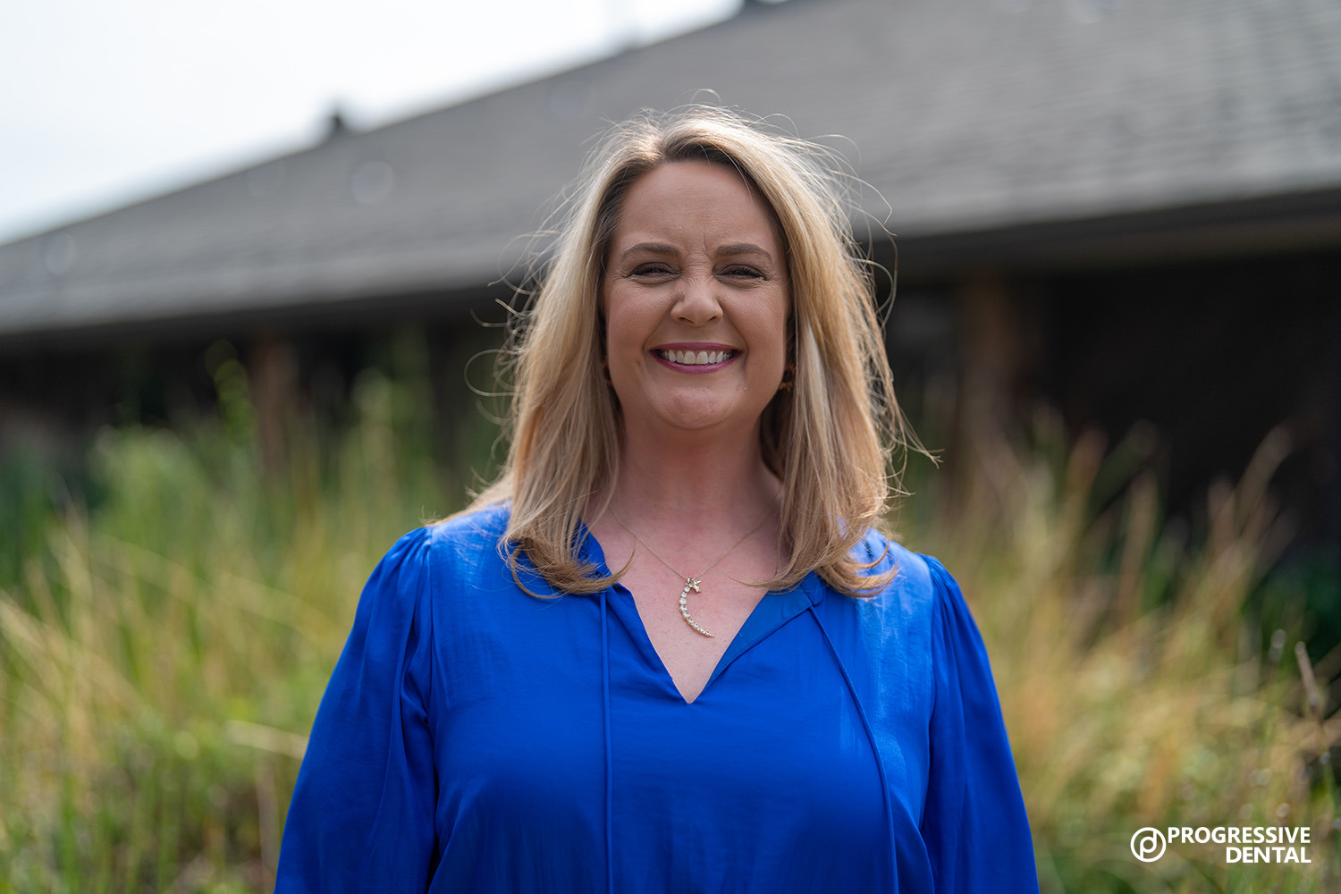 Woman with blonde hair wearing a bright blue blouse smiles outdoors, her confidence shining like the best cosmetic dentist in Oklahoma City could provide.