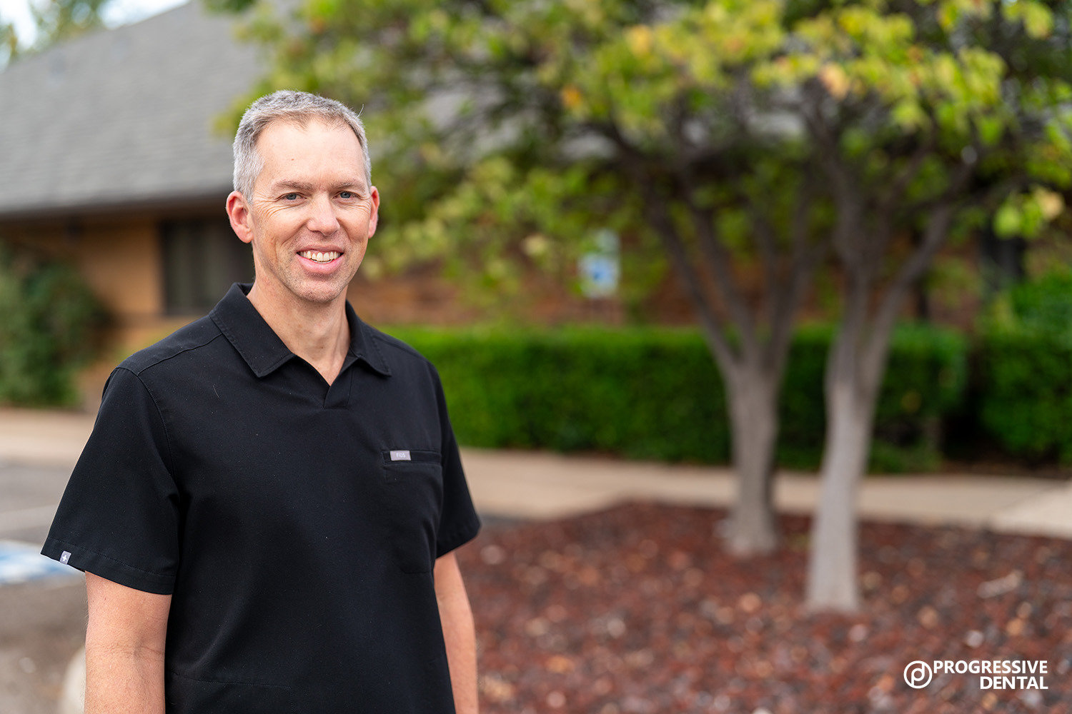 A man in a black shirt stands outside near a building and trees. The logo “Progressive Dental,” home of the best cosmetic dentist in Oklahoma City, appears in the bottom right corner.