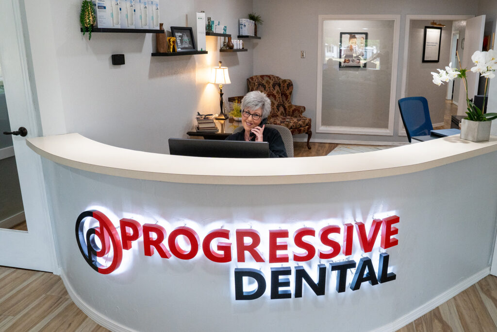 Receptionist on the phone behind a curved front desk with a “Progressive Dental” sign, welcoming dentist without insurance Oklahoma City patients in a modern dental office lobby.