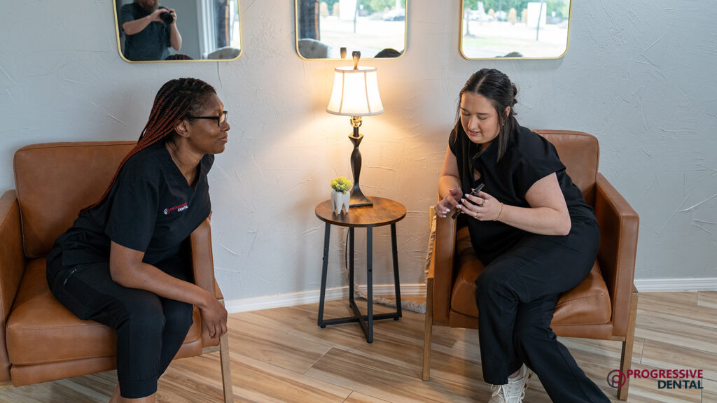 Two Oklahoma City dentist staff members in black uniforms sit in leather chairs, looking at a smartphone, with a small lamp and side table between them.