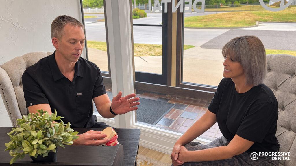 A man holds a dental model and gestures while explaining what to expect during a first dental visit in Oklahoma City to a woman seated across from him inside a dental office.