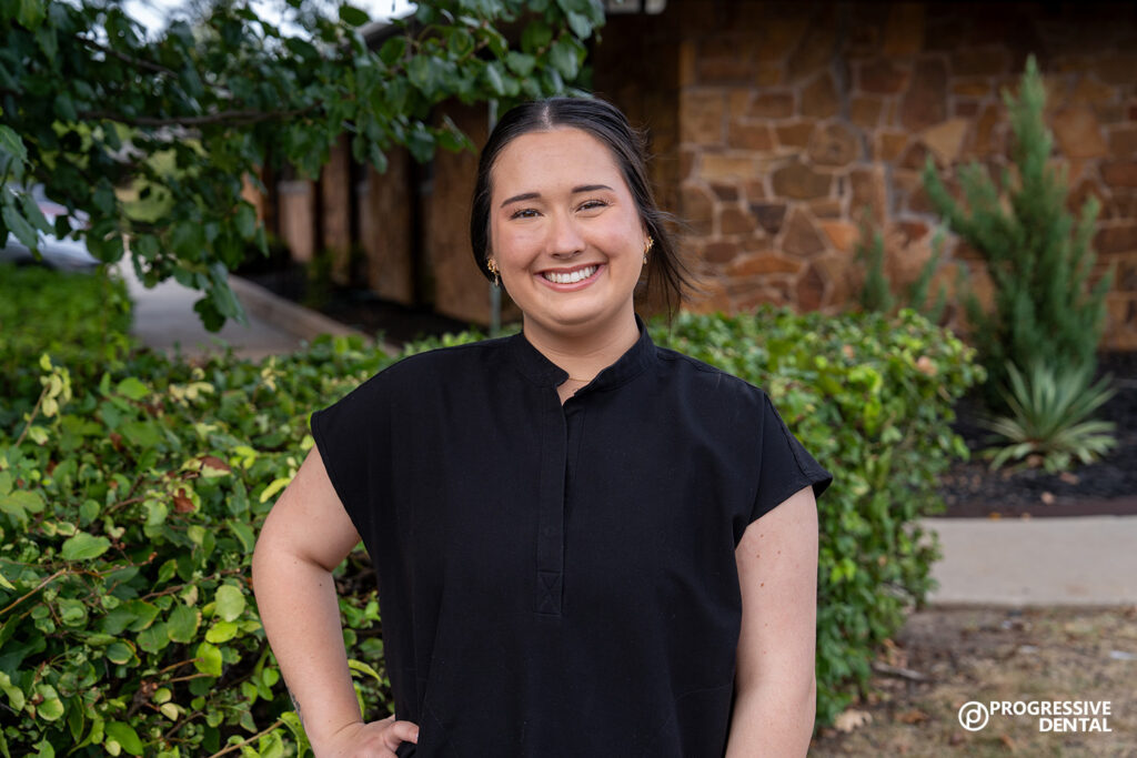 A woman wearing a black shirt stands outside in front of greenery and a stone building, smiling at the camera after her dental exam and cleaning in Oklahoma.