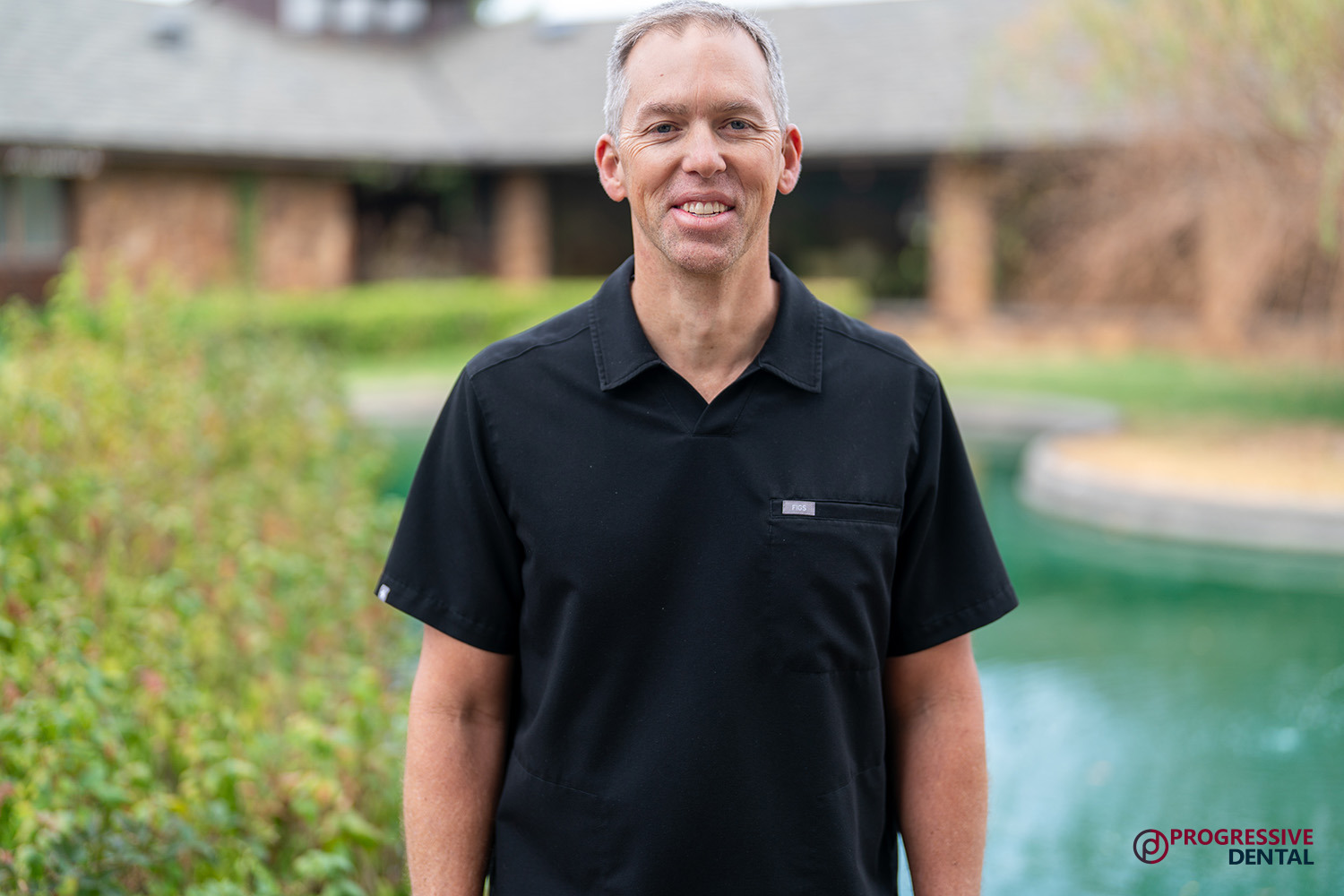 An Oklahoma City cosmetic dentist smiles while wearing a black scrub top outdoors near a pond.